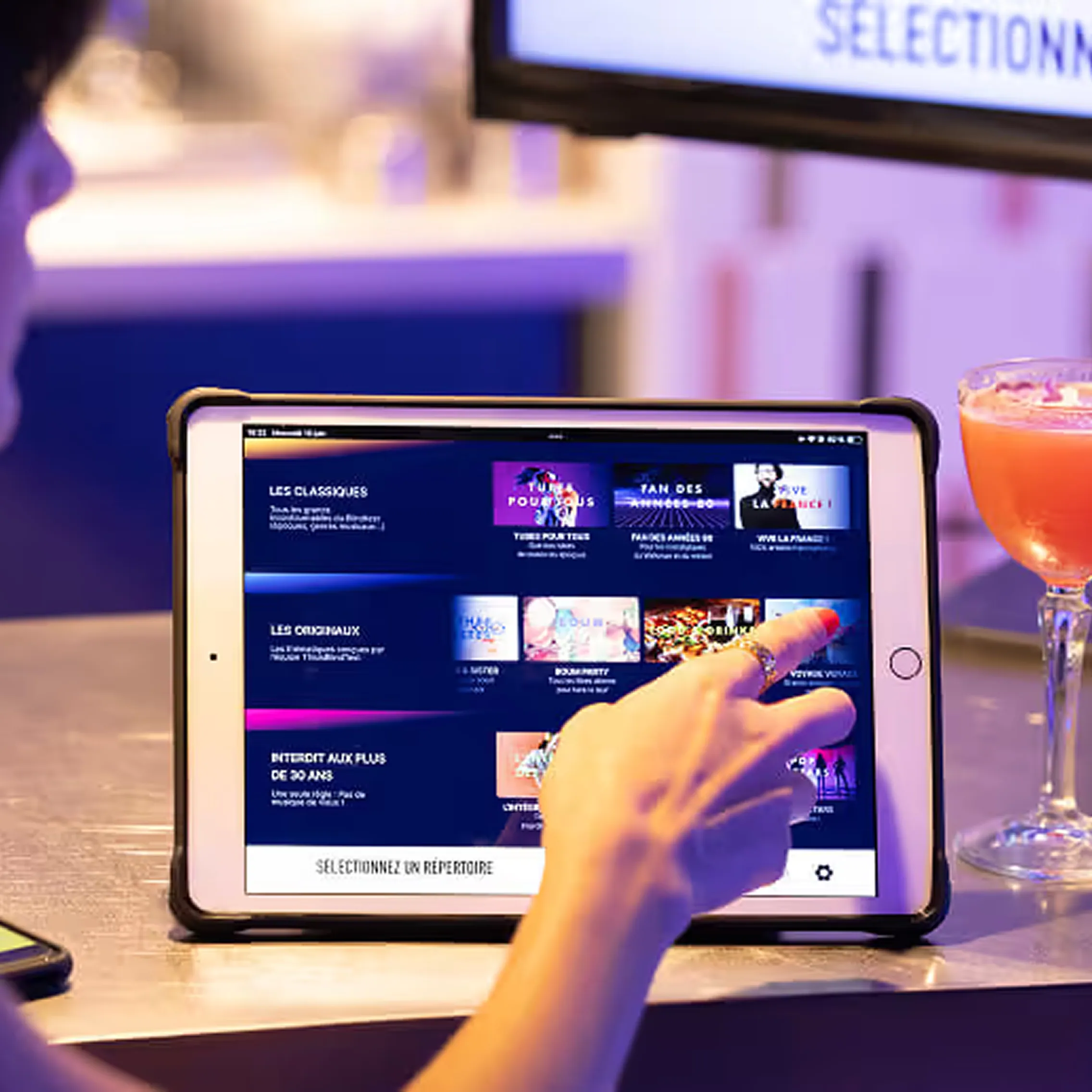Une main sélectionne un répertoire musical sur l’iPad contrôleur, posé sur le comptoir d’un bar